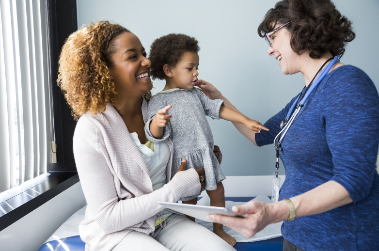 A smiling white female clinician with a stethoscope reaches out to help a smiling black woman and her toddler that is standing on her lap