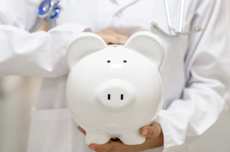 A physician wearing a white coat and a stethoscope holds a white piggy bank in the center of the frame. Only the physician's torso and arms are in the frame.