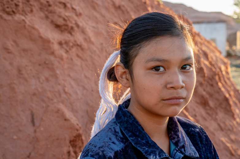 A teenage Native American girl stares seriously into the camera in front of a large, red rock.